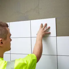 En man i gul väst kaklar en vägg. Han trycker försiktigt en vit kakelplatta mot den spacklade ytan, redo att skapa ett snyggt mönster på våtrumsmatta.