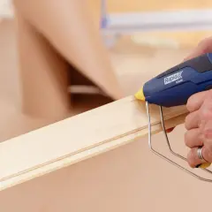 Här limmas golvlister med en limpistol. En person applicerar smältlim på en trälist för att fästa den.