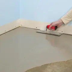 En hand spacklar golv med en bredspackel. Det nya golvet är jämnt och grått.