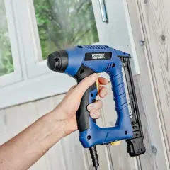 Hand håller en blå kompressor spikpistol mot en trävägg.