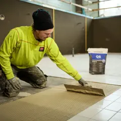Man i gul jacka och mössa sprider ut fix med en tandspackel för att byta golv. En säck med fix står i bakgrunden.
