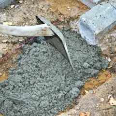 En hacka sprider jordfuktig betong i en grop. Betongblock ligger bredvid.