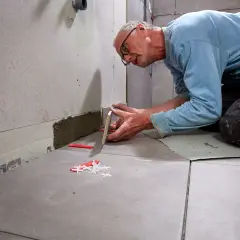 Man limma kakel på väggen, noggrant placerandes en ny platta. Kakelplugg och distanser syns på golvet.