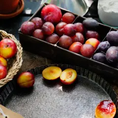 En korg och en låda fyllda med plommon, både mörka och några gula plommon. Halverade plommon ligger på en plåt.