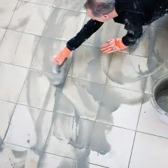 Man med orange handskar arbetar med att måla fogar på ett kaklat golv. En hink med fogmassa står bredvid.