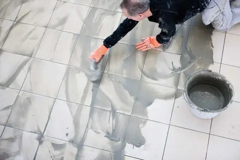 Man med orange handskar arbetar med att måla fogar på ett kaklat golv. En hink med fogmassa står bredvid.