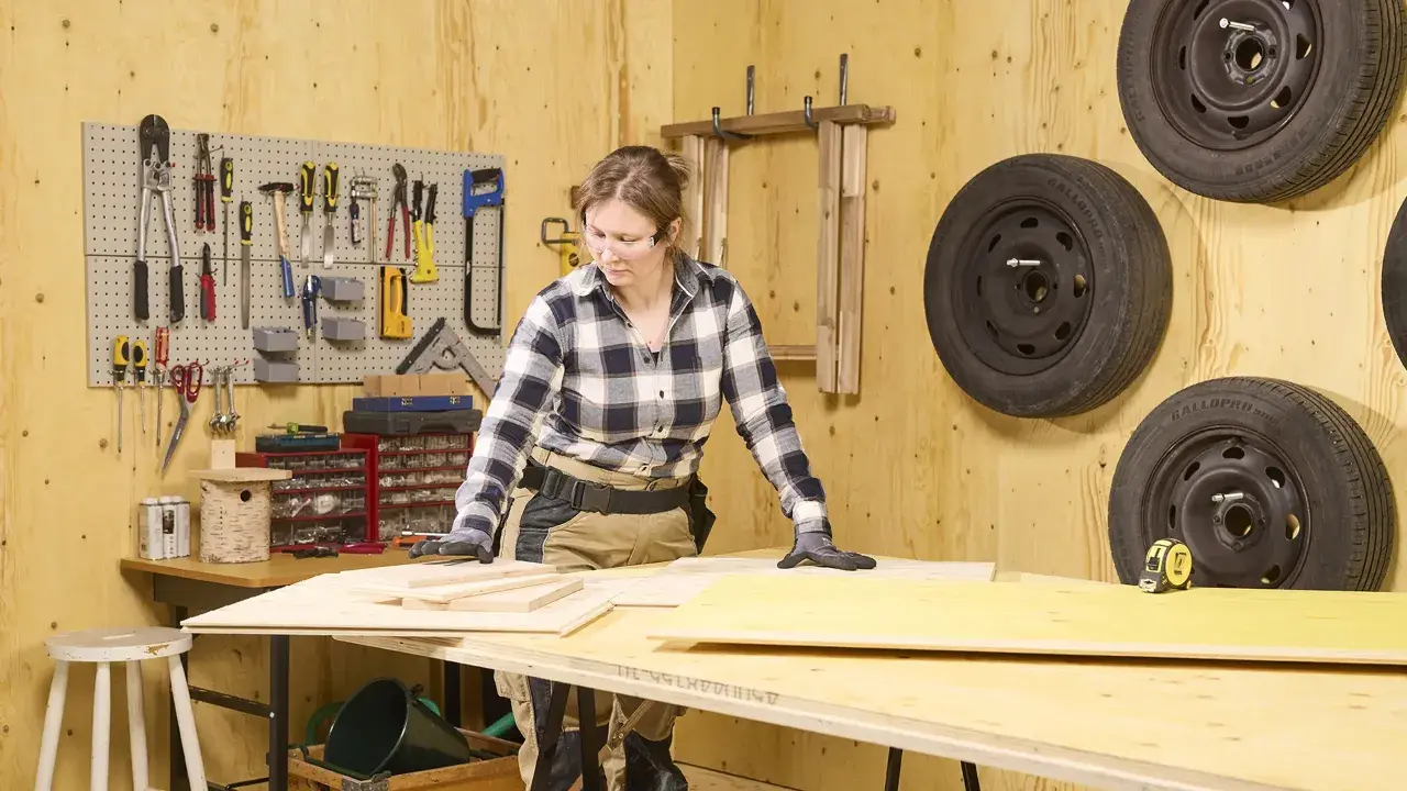 En kvinna arbetar med plywood utomhus i en verkstad. Verktyg hänger på väggen, och däck är upphängda.
