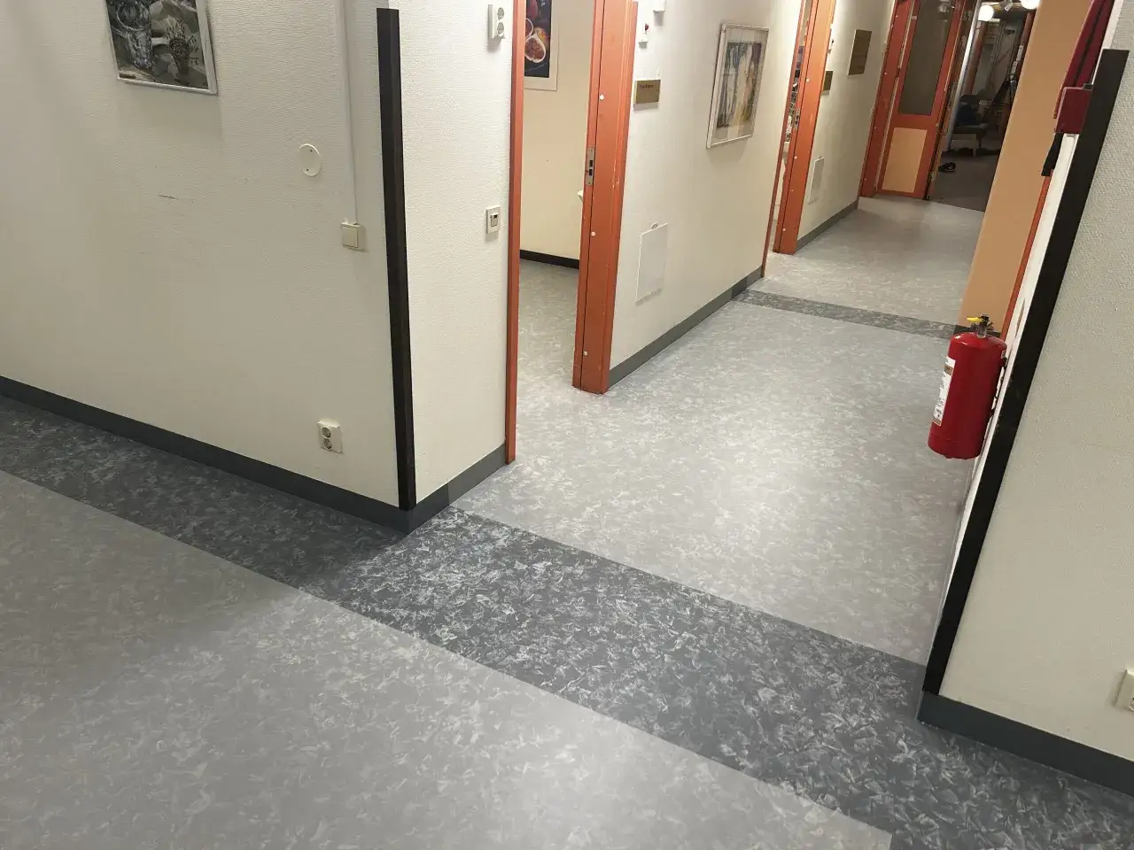 Korridor med ljusgrå golvmatta och mörkare grå rand. Orange dörrkarmar och en röd brandsläckare.