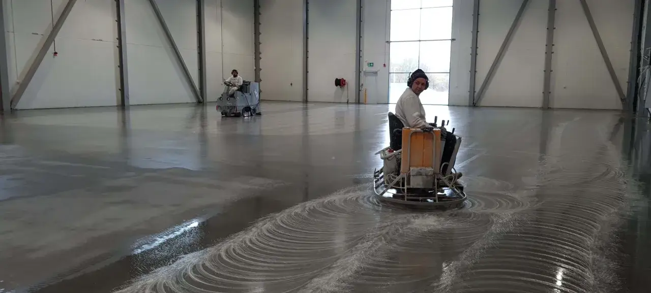 Två personer slipar betonggolv i en stor lokal med maskiner.
