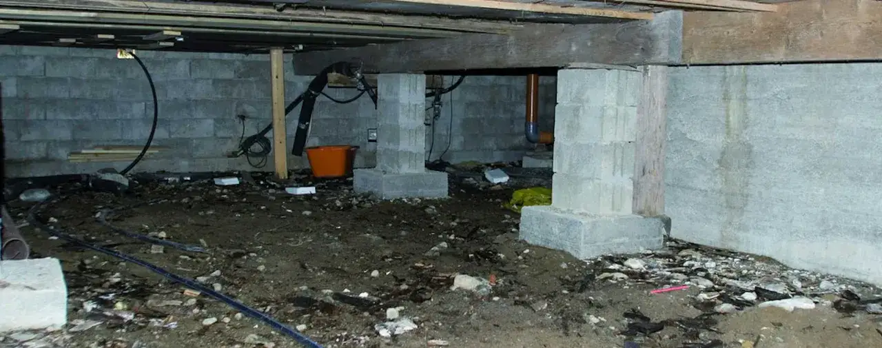 Synlig fukt i krypgrund med betongblock, rör och en orange hink. Marken är fuktig och ojämn.