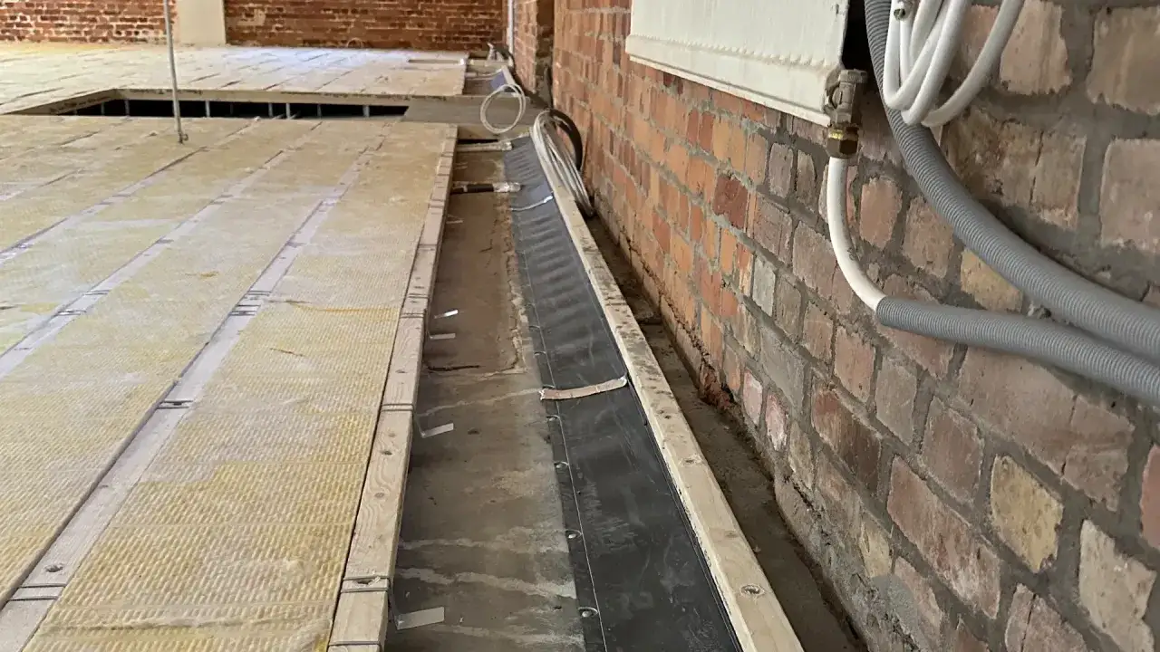 Isolering och träreglar för ett passivt ventilerat golv. En svart plastduk löper längs väggen, redo för installation.