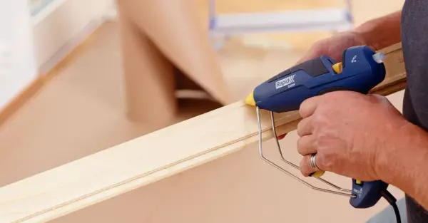 Här limmas golvlister med en limpistol. En person applicerar smältlim på en trälist för att fästa den.